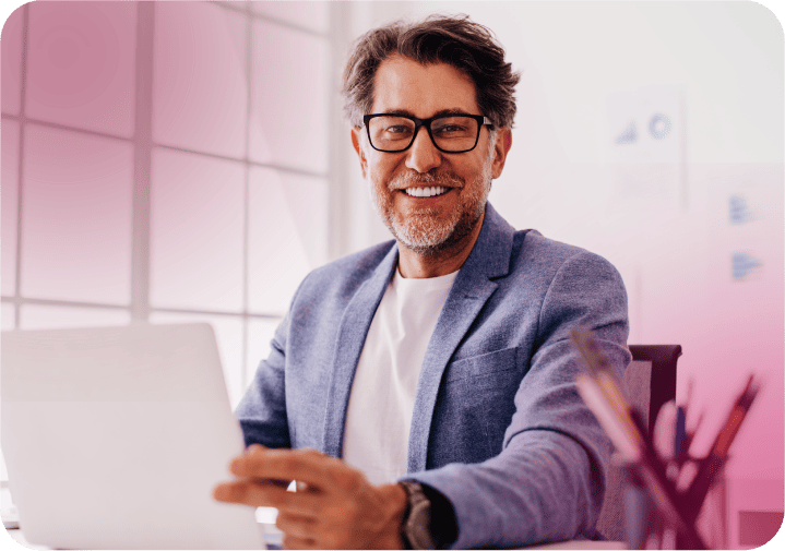 Homem sorridente usando óculos e blazer azul, trabalhando em notebook em ambiente iluminado.  